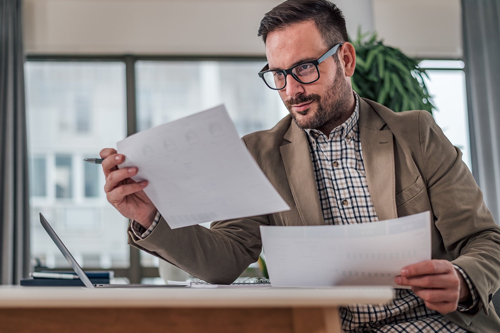 Focused man wearing glasses and a blazer, reviewing documents at a desk with a laptop in a bright office setting.