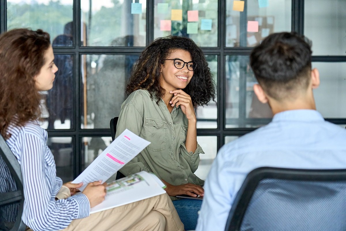 Young woman smiling and engaging in a group discussion with colleagues in a modern office, with notes on the glass wall behind them.