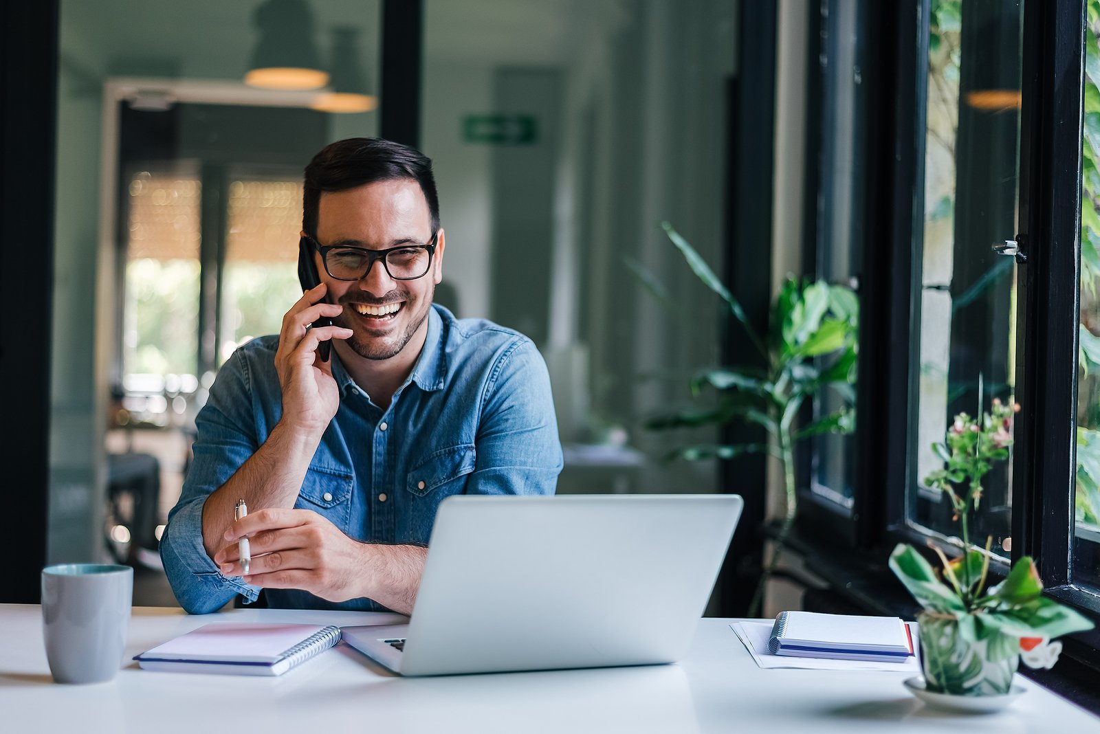 Smiling man wearing glasses and a denim shirt, talking on a smartphone while working on a laptop at a bright, modern office desk with notebooks, a coffee mug, and potted plants.